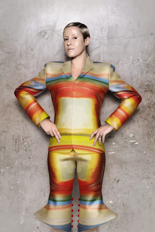Olivia Merquior in a printed blazer and shorts with earth-tone rainbow bands, standing against a concrete wall